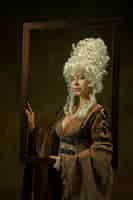 Gratis foto kalmte. portret van middeleeuwse jonge vrouw in vintage kleding met houten frame op donkere achtergrond. vrouwelijk model als hertogin, koninklijk persoon. concept vergelijking van tijdperken, modern, mode, schoonheid.