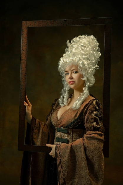 Kalmte. Portret van middeleeuwse jonge vrouw in vintage kleding met houten frame op donkere achtergrond. Vrouwelijk model als hertogin, koninklijk persoon. Concept vergelijking van tijdperken, modern, mode, schoonheid.