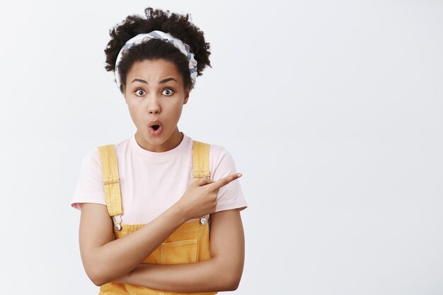 Kan je dat doen. Portret van onder de indruk opgewonden knappe Afro-Amerikaan in gele tuinbroek en hoofdband, naar rechts wijzend, lippen vouwend en opgewonden starend, verbaasd over grijze muur