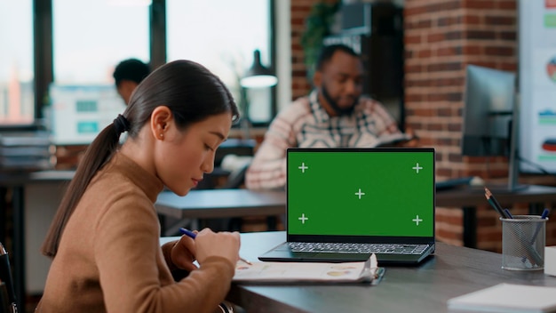 Kantoormedewerker analyseert grafieken op papier en kijkt naar greenscreen op laptop, met behulp van chromakey mockup-sjabloon en geïsoleerde copyspace met lege achtergrond. Vrouw met een handicap.