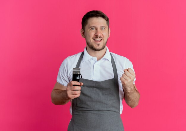 Kapper man in schort bedrijf baard trimmer balde vuist blij en opgewonden staande over roze achtergrond