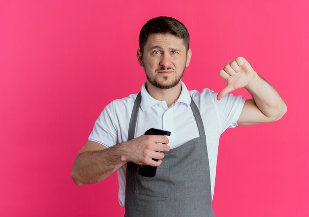 Kapper man in schort bedrijf baard trimmer duimen omlaag wordt ontevreden staande over roze achtergrond te houden