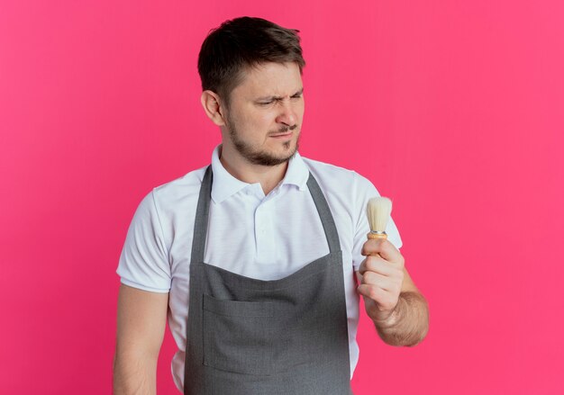 kapper man in schort kijken naar scheerkwast in zijn hand met ernstig gezicht staande over roze muur