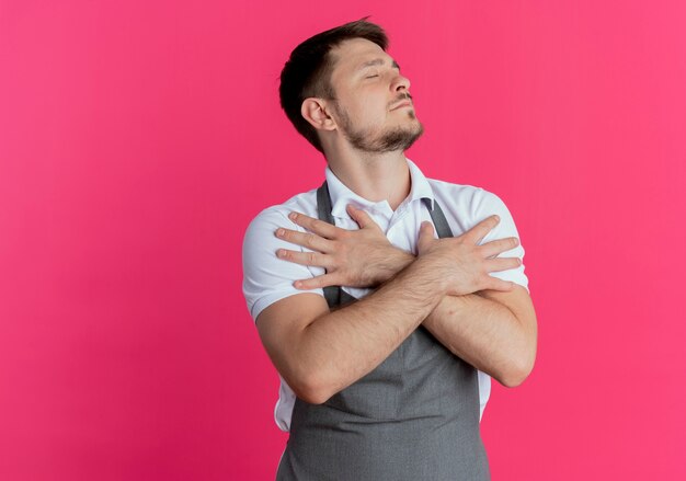 Kapper man in schort met gekruiste armen op zijn borst dankbaar gevoel met gesloten ogen staande over roze achtergrond