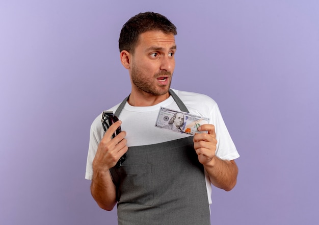 Kapper man in schort met haar snijmachine en contant geld opzij kijken verward staande over paarse muur