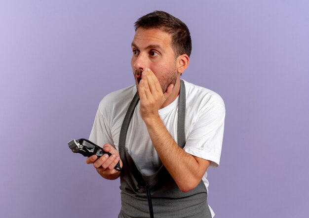 Kapper man in schort met haar snijmachine kijken verbaasd hand in de buurt van mond vertellen een geheim staande over paarse muur