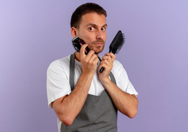 Kapper man in schort met haarsnijmachine en haarborstel naar voren kijkend verward staande over paarse muur