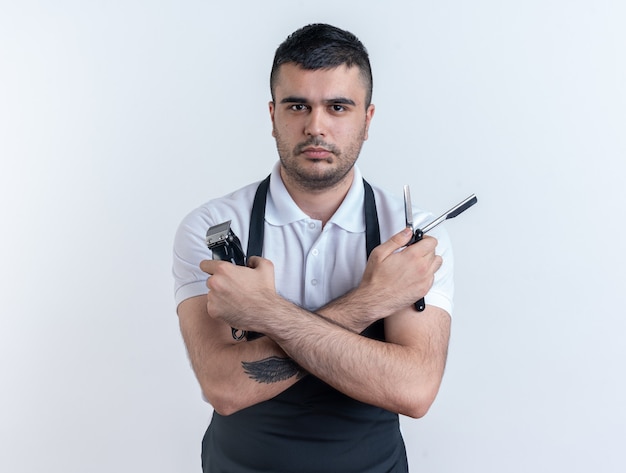 Kapper man in schort met kappers tools kijken camera met ernstige zelfverzekerde uitdrukking staande op witte achtergrond