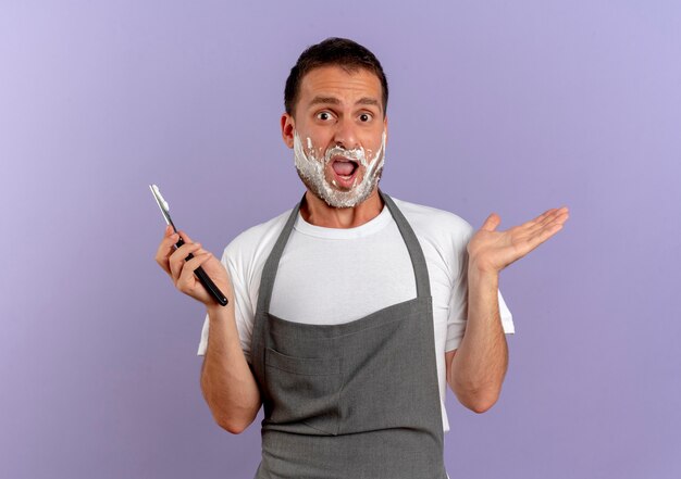 Kapper man in schort met scheerschuim op zijn gezicht met scheermes naar voren kijkend verward schouderophalend staande over paarse muur