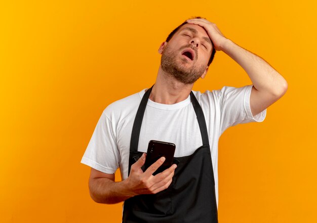 Kapper man in schort met smartphone met hand op zijn hoofd op zoek gehinderd en overwerkt staande over oranje muur