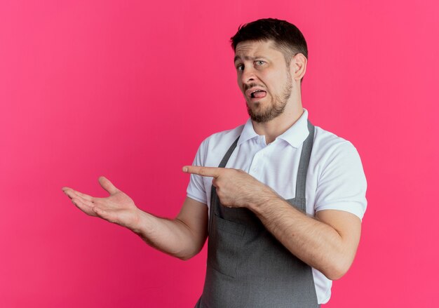 Kapper man in schort presenteren met arm van de hand iets wijst met wijsvinger naar de zijkant staande over roze muur