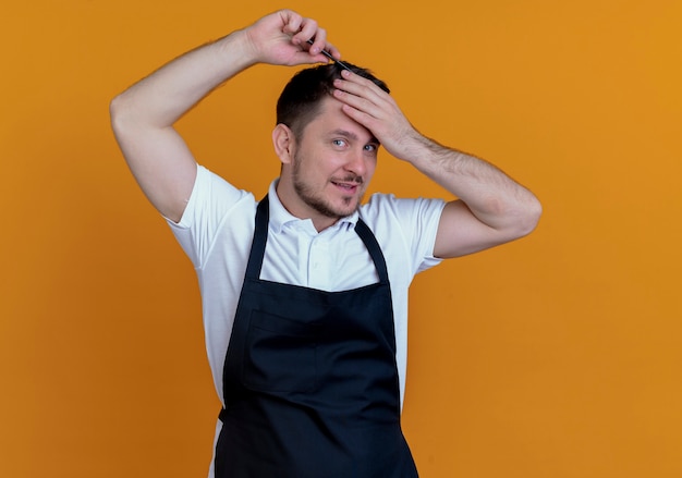 Kapper man in schort zijn haar kammen camera kijken met glimlach op gezicht staande over oranje achtergrond