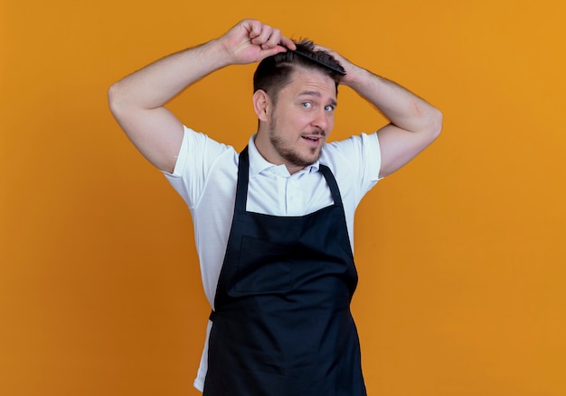 Kapper man in schort zijn haar kammen op zoek zelfverzekerd staande over oranje achtergrond