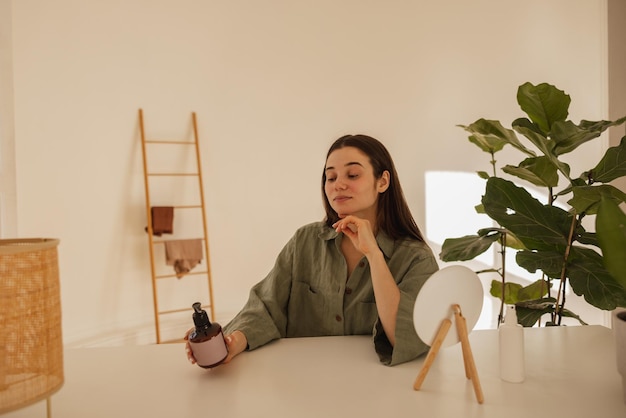 Kaukasisch jong meisje leest zorgvuldig de samenstelling van handcrème op pot terwijl ze binnenshuis aan tafel zit Brunette in groen shirt overtuigd van natuurlijkheid van product Zorgconcept