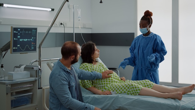 Kaukasisch paar verwacht een baby in de kraamafdeling in het ziekenhuis. Zwangere vrouw zittend in bed praten met Afro-Amerikaanse verpleegster en jonge echtgenoot. Medische hulp bij bevalling