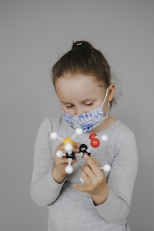 Kaukasisch vrouwelijk kind met een beschermend medisch gezichtsmasker dat speelt met het coronavirus-celmodel