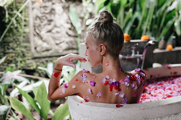 Kaukasisch vrouwelijk model rusten in bad vol bloemblaadjes. spectaculaire jongedame doet spa in de ochtend.