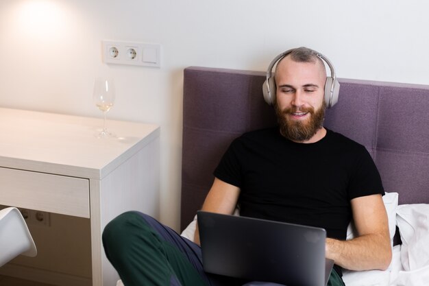 Gratis foto kaukasische bebaarde man in koptelefoon in slaapkamer op bed bezig met laptop vanuit huis, typen, denken.