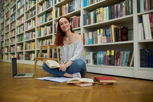 Kennis. Jonge lachende langharige vrouw zittend op parketvloer in bibliotheek met laptop en boeken schrijven in beurt studeren