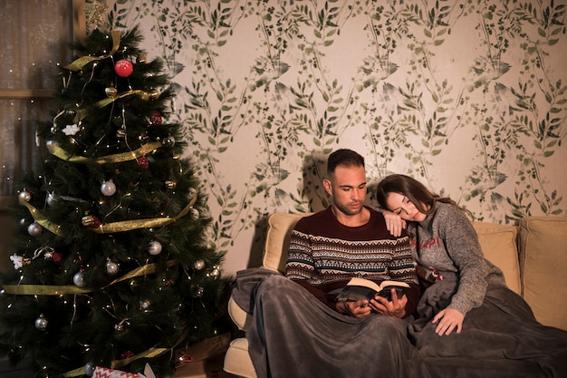 Kerel dichtbij dame in sprei met boek dichtbij Kerstboom