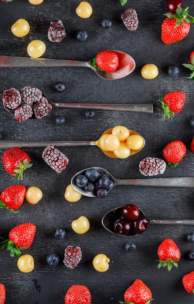 Gratis foto kersen in lepels met aardbeien, bosbessen, moerbeien plat lag op een donkere tafel