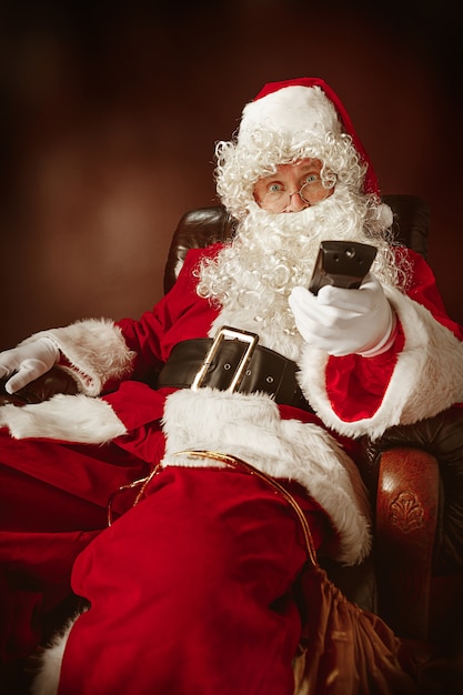 Gratis foto kerstman met een luxe witte baard, kerstmuts en een rood kostuum zittend in een stoel met afstandsbediening van de tv