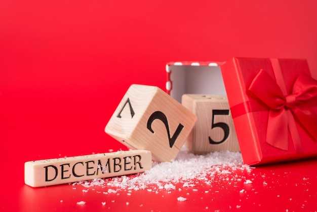 Kerstmis en presenteert concept. close-up foto van houten kubus kalender met datum van kerstmis en een open rode geschenkdoos met rood lint geïsoleerd op rode achtergrond met sneeuw