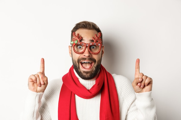 Kerstmis, Nieuwjaar en viering concept. Close-up van knappe bebaarde man in feestbril en rode sjaal, vier wintervakanties en wijzende vingers naar kopieerruimte