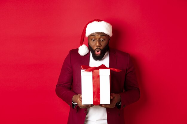 Kerstmis, Nieuwjaar en winkelconcept. Verrast zwarte man die naar kerstcadeau staart, wow verbaasd zegt, vakantiecadeau ontvangt, rode achtergrond