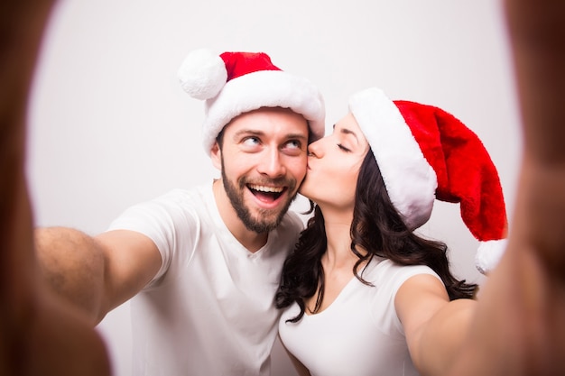 Kerstmis, vakantie, technologie en mensen concept - gelukkige paar in santa hoeden selfie foto te nemen uit handen op witte achtergrond. ze kijken naar de camera en kussen