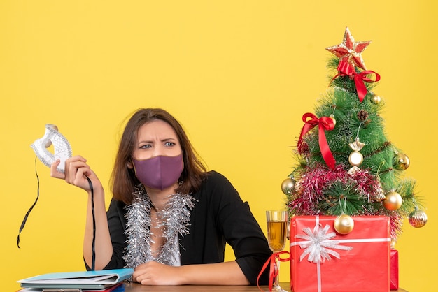 Kerstsfeer met doordachte mooie dame in pak met haar medische masker op kantoor