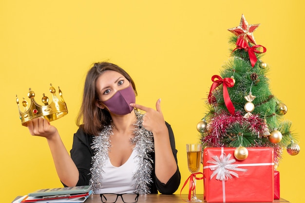 Kerstsfeer met doordachte mooie dame in pak met kroon dragen van haar medische masker in het kantoor