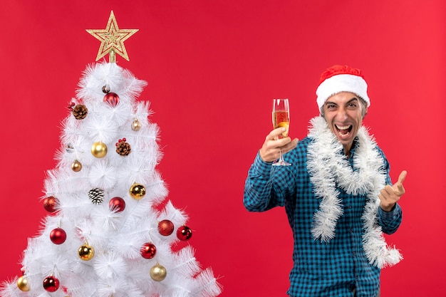 kerstsfeer met emotionele gekke jonge man met kerstman hoed in een blauw gestript shirt met een glas wijn in de buurt van de kerstboom