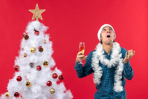 kerstsfeer met emotionele jonge man met kerstman hoed in een blauw gestript shirt met een glas wijn opzoeken in de buurt van de kerstboom