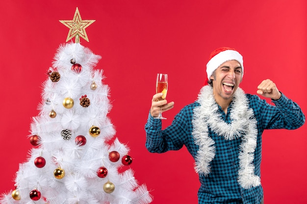 kerstsfeer met emotionele trotse gekke jonge man met kerstman hoed in een blauw gestript shirt met een glas wijn in de buurt van de kerstboom