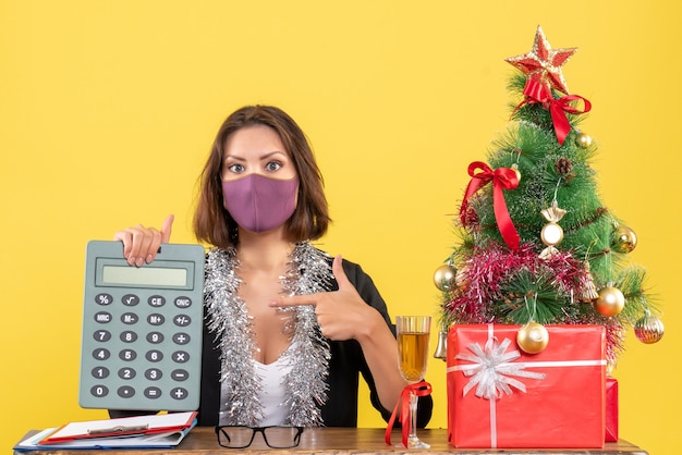 Kerstsfeer met geconcentreerde mooie dame in pak met medische masker wijzende rekenmachine in het kantoor op geel