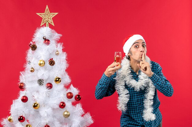 Kerstsfeer met gelukkige gekke emotionele jonge man met kerstman hoed in een blauw gestript shirt een glas wijn heffen en stilte gebaar maken in de buurt van de kerstboom