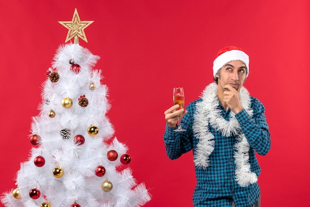 Kerstsfeer met gelukkige gekke emotionele verwarde jonge man met kerstman hoed in een blauw gestript shirt een glas wijn opheffen in de buurt van de kerstboom