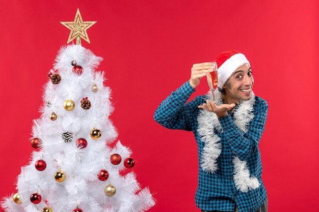 kerstsfeer met gelukkige jonge man met kerstman hoed in een blauw gestript shirt een glas wijn te verhogen in de buurt van de kerstboom