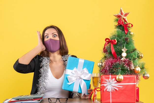 Kerstsfeer met geschokte mooie dame in pak met medisch masker en geschenk te houden op kantoor