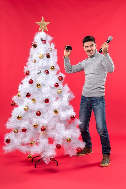 Kerstsfeer met jonge kerel die zich dichtbij versierde kerstboom bevindt en microfoon houdt die foto neemt en trots op zichzelf