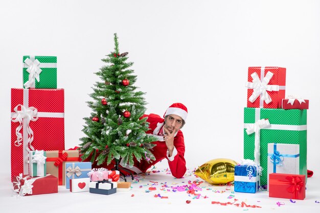 Kerstsfeer met jonge kerstman verstopt achter de kerstboom in de buurt van geschenken in verschillende kleuren op een witte achtergrond