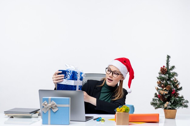 Kerstsfeer met jonge vrouw met kerstman hoed en het dragen van bril zittend aan een tafel met haar cadeau op witte achtergrond