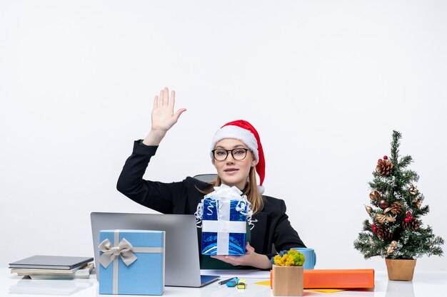 Kerstsfeer met jonge vrouw met kerstman hoed en het dragen van een bril zittend aan een tafel met cadeau en hallo zeggen tegen iemand op witte achtergrond