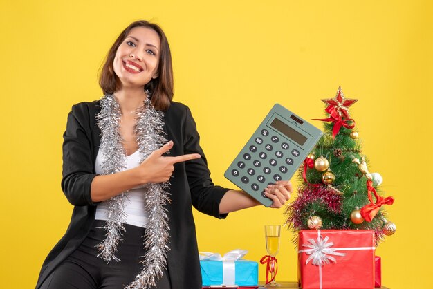 Kerstsfeer met lachende mooie dame permanent in het kantoor en rekenmachine op kantoor op geel te wijzen