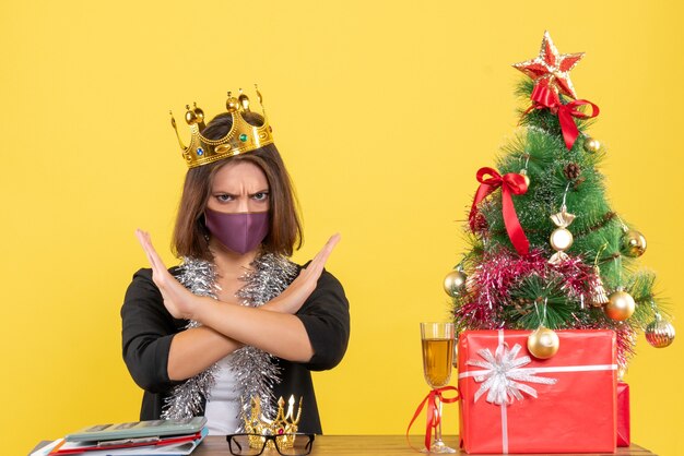 Kerstsfeer met mooie dame in pak met medisch masker en het dragen van masker negatief gebaar maken in het kantoor op geel