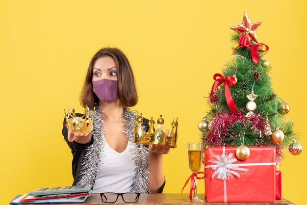 Kerstsfeer met mooie dame in pak met medisch masker en kronen in het kantoor op geel te houden