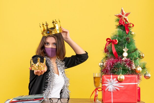Kerstsfeer met mooie dame in pak met medisch masker en kronen op het hoofd en in de hand in het kantoor op geel