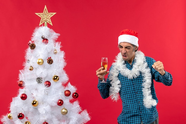 Kerstsfeer met trotse jongeman met kerstman hoed in een blauw gestript shirt die een glas wijn opheft en zijn geluk toont in de buurt van de kerstboom