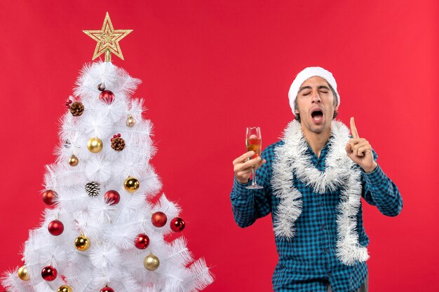 kerstsfeer met zelfverzekerde vastberaden emotionele jongeman met kerstman hoed in een blauw gestript shirt een glas wijn verhogen in de buurt van de kerstboom